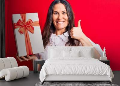 Young beautiful brunette woman holding birthday gift standing over isolated red background smiling happy and positive, thumb up doing excellent and approval sign Wall mural