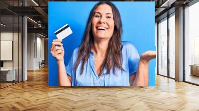 Young beautiful brunette woman holding credit card over isolated blue background celebrating achievement with happy smile and winner expression with raised hand Wall mural