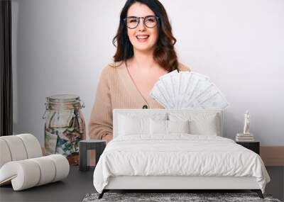 Young beautiful brunette woman holding jar with savings holding dollars looking positive and happy standing and smiling with a confident smile showing teeth Wall mural