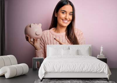 Young beautiful brunette woman holding piggy bank saving money for retirement very happy pointing with hand and finger Wall mural