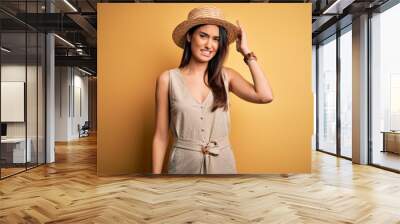 Young beautiful brunette woman on vacation wearing casual dress and hat confuse and wonder about question. Uncertain with doubt, thinking with hand on head. Pensive concept. Wall mural