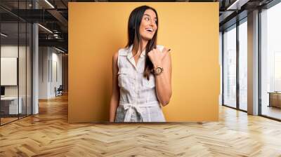 Young beautiful brunette woman on vacation wearing casual dress over yellow background smiling with happy face looking and pointing to the side with thumb up. Wall mural