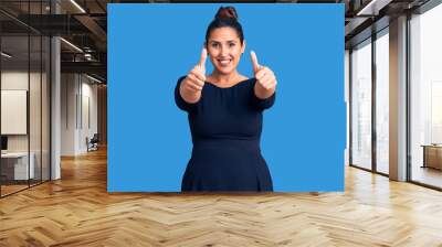 Young beautiful brunette woman wearing casual dress approving doing positive gesture with hand, thumbs up smiling and happy for success. winner gesture. Wall mural