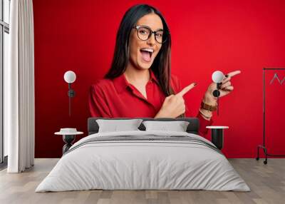 Young beautiful brunette woman wearing casual shirt and glasses over red background smiling and looking at the camera pointing with two hands and fingers to the side. Wall mural