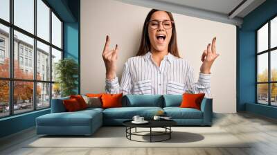 Young beautiful brunette woman wearing casual shirt and glasses over white background shouting with crazy expression doing rock symbol with hands up. Music star. Heavy concept. Wall mural