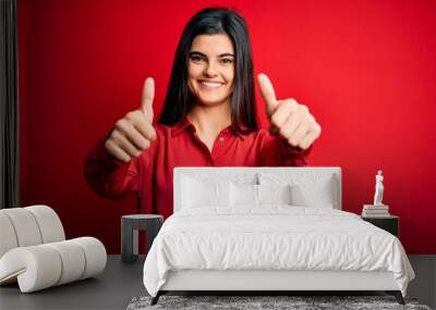 Young beautiful brunette woman wearing casual shirt standing over red background approving doing positive gesture with hand, thumbs up smiling and happy for success. Winner gesture. Wall mural