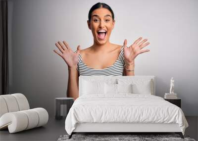 Young beautiful brunette woman wearing casual striped dress over isolated white background celebrating crazy and amazed for success with arms raised and open eyes screaming excited. Winner concept Wall mural