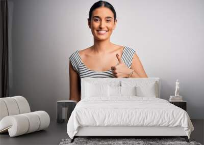 Young beautiful brunette woman wearing casual striped dress over isolated white background doing happy thumbs up gesture with hand. Approving expression looking at the camera showing success. Wall mural