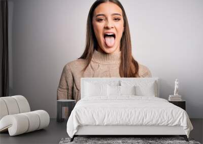 Young beautiful brunette woman wearing casual sweater standing over white background sticking tongue out happy with funny expression. Emotion concept. Wall mural