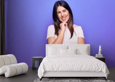 Young beautiful brunette woman wearing casual t-shirt standing over purple background smiling looking confident at the camera with crossed arms and hand on chin. Thinking positive. Wall mural