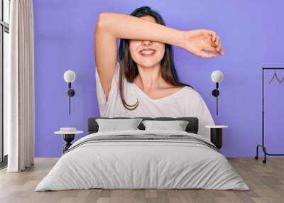 Young beautiful brunette woman wearing casual white t-shirt over purple background covering eyes with arm smiling cheerful and funny. Blind concept. Wall mural