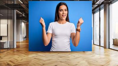 Young beautiful brunette woman wearing white casual t-shirt standing over blue background celebrating surprised and amazed for success with arms raised and open eyes. Winner concept. Wall mural