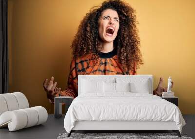 Young beautiful brunette woman with curly hair and piercing wearing casual t-shirt crazy and mad shouting and yelling with aggressive expression and arms raised. Frustration concept. Wall mural