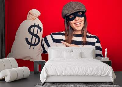 Young beautiful burglar woman wearing cap and mask holding money bag with dollar symbol smiling happy pointing with hand and finger Wall mural