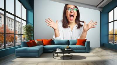 Young beautiful business woman wearing glasses over isolated background celebrating crazy and amazed for success with arms raised and open eyes screaming excited. Winner concept Wall mural