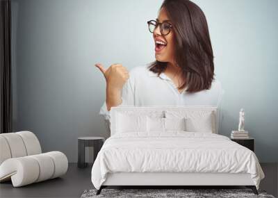 Young beautiful business woman wearing glasses over isolated background smiling with happy face looking and pointing to the side with thumb up. Wall mural