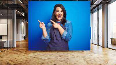 Young beautiful business woman wearing store uniform apron over blue isolated background smiling and looking at the camera pointing with two hands and fingers to the side. Wall mural