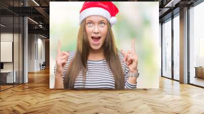 Young beautiful caucasian woman wearing christmas hat over isolated background amazed and surprised looking up and pointing with fingers and raised arms. Wall mural