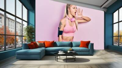 Young beautiful caucasian woman wearing gym clothes and using headphones shouting and screaming loud to side with hand on mouth. communication concept. Wall mural