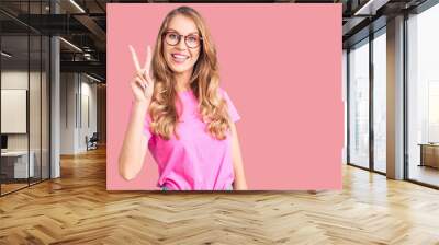 Young beautiful caucasian woman with blond hair wearing casual clothes and glasses showing and pointing up with fingers number two while smiling confident and happy. Wall mural