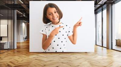 Young beautiful child girl wearing casual t-shirt standing over isolated white background smiling and looking at the camera pointing with two hands and fingers to the side. Wall mural