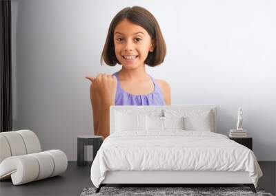 Young beautiful child girl wearing purple casual dress standing over isolated white background smiling with happy face looking and pointing to the side with thumb up. Wall mural