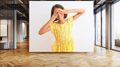 Young beautiful child girl wearing yellow floral dress standing over isolated white background covering eyes with hands smiling cheerful and funny. Blind concept. Wall mural