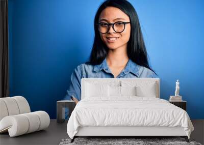 Young beautiful chinese woman wearing casual denim shirt over isolated blue background happy face smiling with crossed arms looking at the camera. Positive person. Wall mural