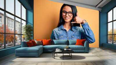 Young beautiful chinese woman wearing casual denim shirt over isolated yellow background smiling doing phone gesture with hand and fingers like talking on the telephone. Communicating concepts. Wall mural