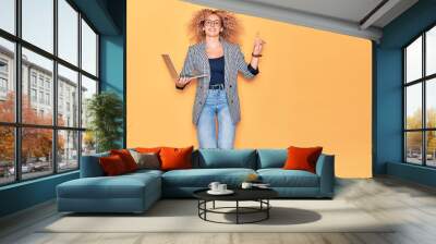 Young beautiful curly businesswoman wearing glasses smiling happy. Jumping with smile on face working using laptop over isolated yellow background. Wall mural