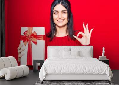 Young beautiful girl holding gift doing ok sign with fingers, smiling friendly gesturing excellent symbol Wall mural