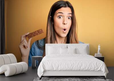 Young beautiful girl holding healthy protein bar standing over isolated yellow background scared in shock with a surprise face, afraid and excited with fear expression Wall mural