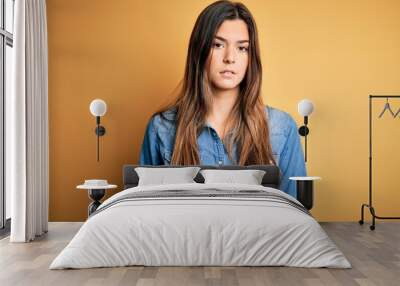 Young beautiful girl wearing casual denim shirt standing over isolated yellow background Relaxed with serious expression on face. Simple and natural looking at the camera. Wall mural