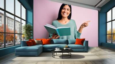 Young beautiful hispanic student woman reading a book over pink isolated background very happy pointing with hand and finger to the side Wall mural