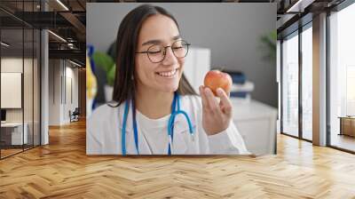 Young beautiful hispanic woman doctor smiling confident holding apple at clinic Wall mural