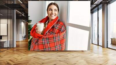 Young beautiful hispanic woman drinking glass of milk standing by christmas tree at home Wall mural