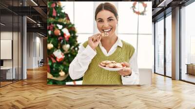 Young beautiful hispanic woman eating cookies standing by christmas tree at home Wall mural