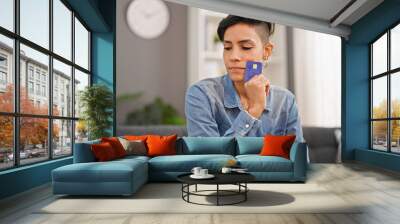 Young beautiful hispanic woman holding credit card sitting on sofa thinking at home Wall mural