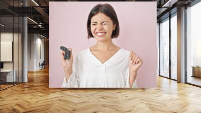 Young beautiful hispanic woman pointing to key of new car with winner expression over isolated pink background Wall mural