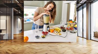 Young beautiful hispanic woman preparing vegetable smoothie with blender at the kitchen Wall mural