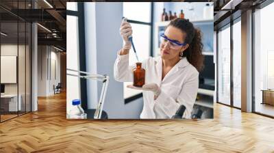 Young beautiful hispanic woman scientist pouring liquid on bottle at laboratory Wall mural