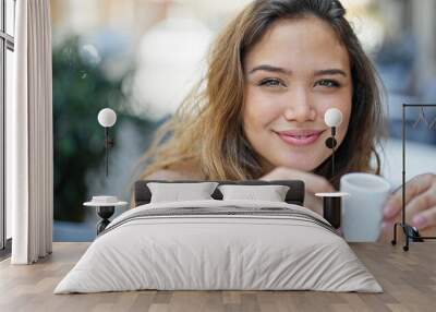 Young beautiful hispanic woman sitting on table drinking coffee smiling at coffee shop terrace Wall mural