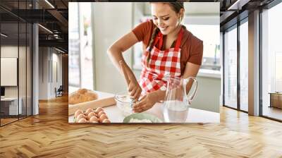 Young beautiful hispanic woman smiling confident mixing water and flour on bowl at the kitchen Wall mural