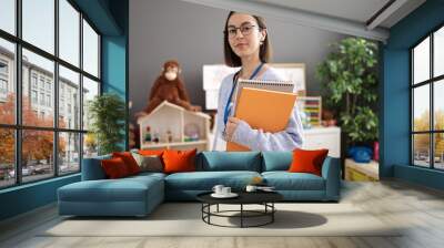 Young beautiful hispanic woman teacher holding books standing at kindergarten Wall mural