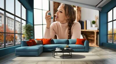 Young beautiful hispanic woman using inhaler sitting on table at home Wall mural