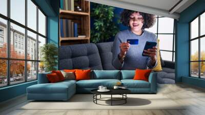 Young beautiful hispanic woman using smartphone and credit card sitting on sofa at home Wall mural