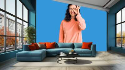 Young beautiful hispanic woman wearing casual clothes doing ok gesture with hand smiling, eye looking through fingers with happy face. Wall mural