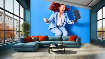 Young beautiful latin businesswoman smiling happy celebrating success. Jumping with smile on face using laptop over isolated blue background Wall mural