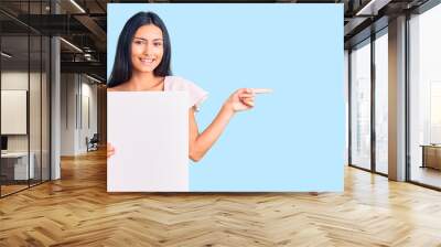 Young beautiful latin girl holding blank empty banner smiling happy pointing with hand and finger to the side Wall mural