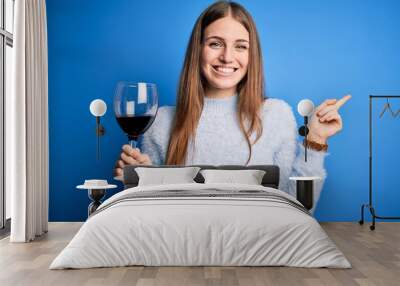 Young beautiful redhead woman drinking glass of red wine over isolated blue background very happy pointing with hand and finger to the side Wall mural